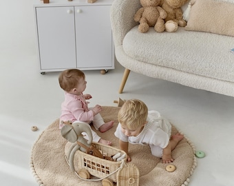 May include: Two toddlers play on a beige round rug with a wooden elephant toy pulling a wicker basket filled with toys. The rug has a white pom-pom trim.