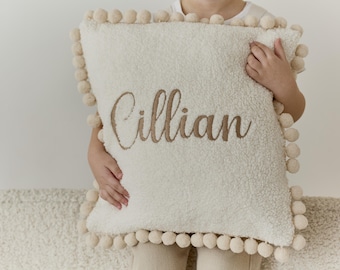 Könnte beinhalten: Ein weißes, flauschiges Kissen mit einem braunen gestickten Namen "Cillian". Das Kissen hat einen weißen Pompon-Besatz.
