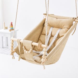 May include: A beige fabric baby swing suspended by thick ropes, featuring a padded seat, a wooden front bar adorned with light-colored wooden beads, and a grey safety harness system. The swing is designed with tie-top attachments for hanging.