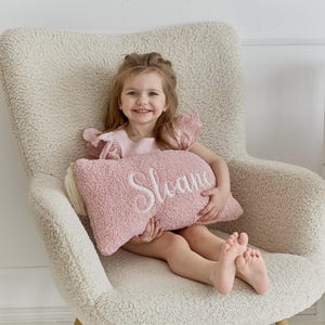 Peut inclure: Une jeune fille est assise dans un fauteuil crème avec un coussin rose sur lequel est inscrit "Sloane". Elle porte une robe rose et a un grand sourire.