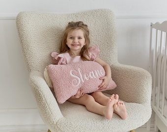 May include: A young girl sits in a cream-colored armchair with a pink pillow that says "Sloane" on it. She is wearing a pink dress and has a big smile on her face.