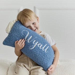 Puede incluir: Una almohada rectangular azul con el nombre "Wyatt" bordado en escritura blanca. La almohada tiene una apariencia texturizada y afelpada y una borla blanca en un extremo. Un niño sostiene la almohada.