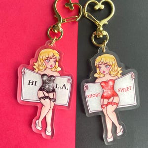 May include: Two clear acrylic keychains featuring cartoon women in lingerie holding signs. The woman on the left holds a sign that says "HI L.A." and the woman on the right holds a sign that says "SHORT SWEET".