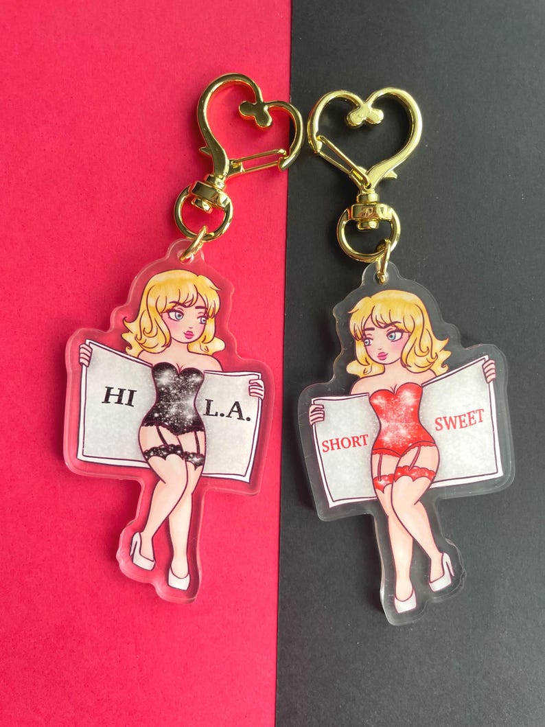 May include: Two clear acrylic keychains featuring cartoon women in lingerie holding signs. The woman on the left holds a sign that says "HI L.A." and the woman on the right holds a sign that says "SHORT SWEET".