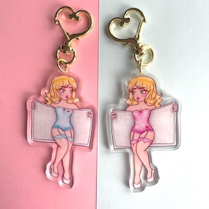 May include: Two keychains featuring cartoon women in lingerie holding blank signs. The women have blonde hair and are wearing blue and pink lingerie. The keychains are made of clear acrylic and have gold heart-shaped clasps.