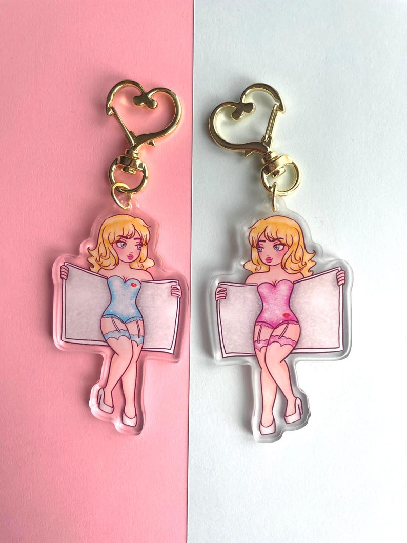 May include: Two keychains featuring cartoon women in lingerie holding blank signs. The women have blonde hair and are wearing blue and pink lingerie. The keychains are made of clear acrylic and have gold heart-shaped clasps.