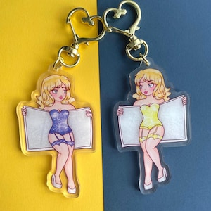 May include: Two clear acrylic keychains featuring cartoon women in lingerie holding blank signs. One woman is wearing a blue and silver sparkly outfit, the other is wearing a yellow outfit. Both have blonde hair and are smiling.