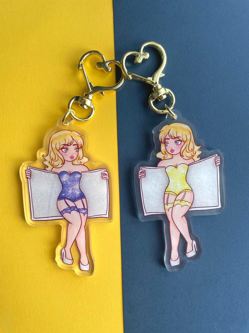 May include: Two clear acrylic keychains featuring cartoon women in lingerie holding blank signs. One woman is wearing a blue and silver sparkly outfit, the other is wearing a yellow outfit. Both have blonde hair and are smiling.