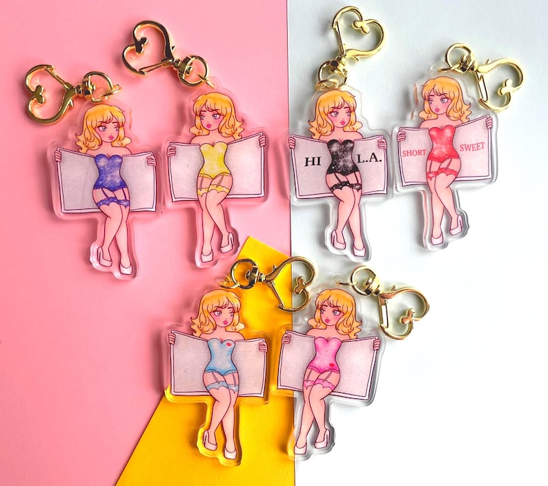 May include: Six colorful acrylic keychains featuring a cartoon woman in lingerie holding a sign. The signs read "HI", "L.A.", "SHORT", "SWEET", and two blank signs.
