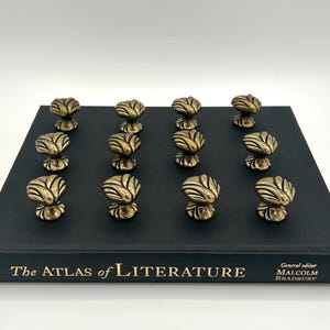May include: Twelve brass-toned metal knobs with a shell-like, ribbed design arranged in a grid on top of a black hardcover book titled The Atlas of Literature, edited by Malcolm Bradbury, featuring gold lettering on the front cover spine.