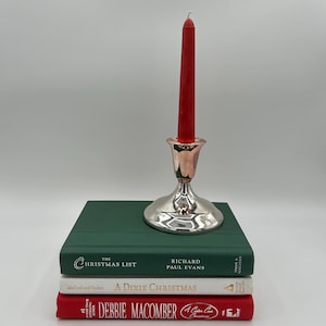 May include: A red taper candle sits in a polished silver candlestick holder, which is placed on a stack of three books. The books are titled The Christmas List by Richard Paul Evans, A Dixie Christmas, and a book by Debbie Macomber.