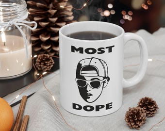 May include: White ceramic mug with the words "MOST DOPE" and a black and white graphic of a person wearing a baseball cap and sunglasses. The mug is filled with a dark liquid.