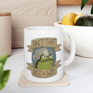 May include: White ceramic mug featuring a cartoon frog with a teapot on its head, set against a blue backdrop. The mug has the text "AIN'T THAT JUST THE WAY" on a banner. The mug is on a square coaster.