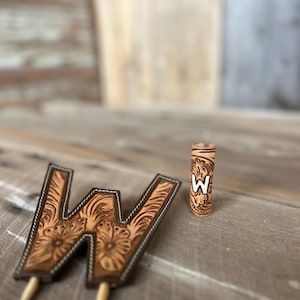 May include: A tan leather topper shaped like the letter W with intricate floral tooling and white stitching, featuring two pointed wooden picks at the base, alongside a matching cylindrical leather sleeve with a white W letter inlay.