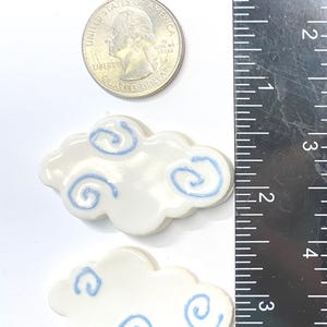 May include: Two white ceramic cloud-shaped ornaments with light blue swirl patterns. A United States quarter coin is placed above the clouds for scale, and a black ruler with white markings shows the items are approximately 2 inches in width.