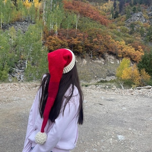 Puede incluir: Un gorro de Papá Noel largo, rojo y blanco con un pompón color crema. El gorro lo lleva una persona de pie al aire libre con un fondo de montaña. El gorro está hecho de material de punto.