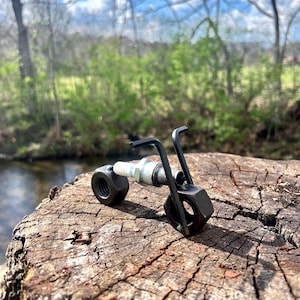 May include: A miniature motorcycle sculpture crafted from metal nuts, a spark plug, and a hex key. The sculpture is set on a weathered tree stump, with a natural backdrop of a stream and foliage. The motorcycle is a unique piece of art.