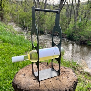 May include: A black metal wine bottle holder with a bottle of white wine inside. The holder is sitting on a tree stump in a grassy area.