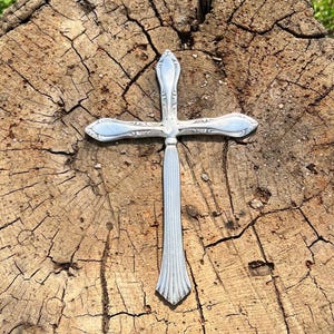 May include: A silver-toned metal cross ornament featuring a vertical base with vertical fluted lines and a horizontal crossbar decorated with ornate, embossed scrollwork patterns at the ends, creating a polished and reflective surface finish.
