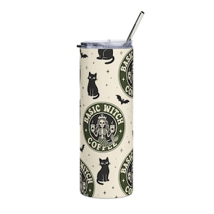 May include: A cylindrical tumbler with a clear lid and a metal straw. The cream-colored surface features a repeating pattern of black cats, bats, small stars, and a circular green logo containing a skeleton mermaid and the text BASIC WITCH COFFEE.