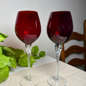 May include: Two wine glasses with deep red bowls and clear, twisted glass stems. Each glass features a rounded, bulbous bowl shape and a circular base, providing a stable foundation for the elegant, spiraled stem design.