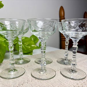 May include: A set of four clear glass stemmed goblets featuring a delicate etched floral and leaf pattern around the rim. Each glass has a multi-faceted, decorative stem and a wide, circular base, providing a stable foundation for the bowl.