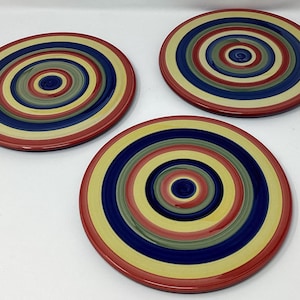 May include: Three round ceramic plates with concentric circles in red, yellow, blue, and green. The plates have a glossy finish and are arranged on a white surface. The plates are decorative and suitable for serving food.