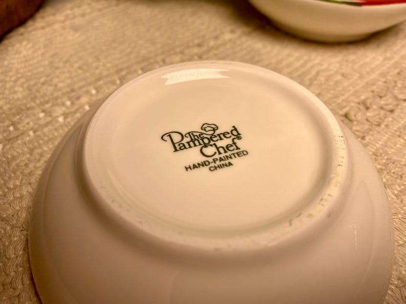 Puede incluir: Un cuenco de cer&aacute;mica blanco con el texto "The Pampered Chef HAND-PAINTED CHINA" impreso en negro. El cuenco es ligeramente curvado y parece nuevo.