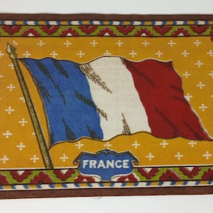 May include: Rectangular decorative mat featuring the French flag in blue, white, and red. The flag is on a yellow background with a repeating cross pattern. The word "FRANCE" is in a blue label at the bottom. The mat has a brown border with floral accents.