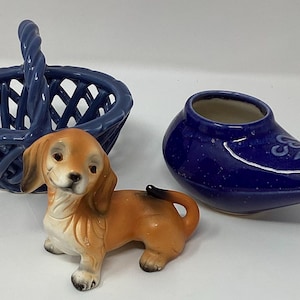 May include: A blue ceramic basket, a brown and tan dachshund figurine, and a blue ceramic neti pot are displayed. The basket has a handle and an open weave design. The neti pot has a spout and a decorative design.