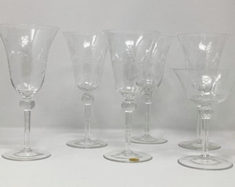 May include: A set of six clear, etched glass goblets and champagne glasses. The glasses have a floral design and are arranged on a white surface. The goblets have a bulbous stem and flared bowl. The champagne glasses have a shallow bowl.