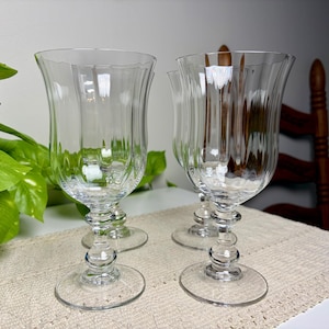 May include: A set of clear glass goblets featuring vertical fluted detailing on the bowl and a decorative multi-tiered stem design. Each glass has a wide, circular base and a flared rim, crafted from transparent glass with a smooth finish.