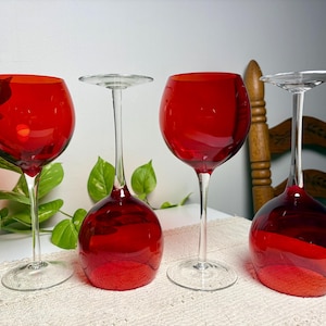May include: Four wine glasses with deep red bowls and clear glass stems and bases. Two glasses are standing upright, while two are inverted, resting on their circular rims. The set features a consistent design with smooth, rounded glass bowls.
