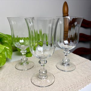 May include: Three clear glass goblets featuring vertical fluted detailing on the bowls, a rounded stem design with a central decorative knob, and a circular base. The glassware is transparent with a smooth finish and a flared rim at the top.