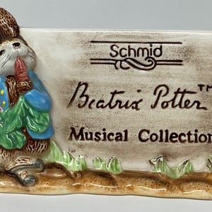 May include: A ceramic figurine featuring Peter Rabbit holding carrots, with a sign that reads "Schmid Beatrix Potter Musical Collection." The figurine has a blue jacket and green leaves, set on a brown base with grass.