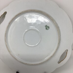 May include: A white porcelain plate with a scalloped edge and two handle cutouts. The underside features a central ring, a small green emblem, and a gold-colored rim. The plate is supported by a black stand.