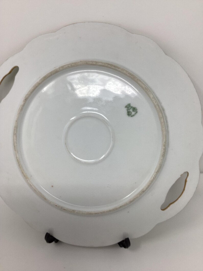 May include: A white porcelain plate with a scalloped edge and two handle cutouts. The underside features a central ring, a small green emblem, and a gold-colored rim. The plate is supported by a black stand.