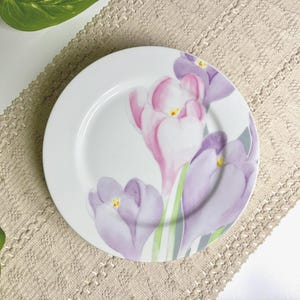 May include: A round white ceramic plate featuring a floral design of three crocus flowers in shades of light pink and purple with green stems and yellow centers. The plate has a smooth, glossy finish and a simple, clean edge design.