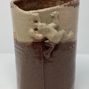 May include: A handmade ceramic tumbler with a two-tone design. The upper portion is a speckled tan, while the lower half is a rich brown. A detailed frog sculpture adorns the side. The tumbler is approximately 6 inches tall.