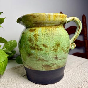 May include: A ceramic pitcher featuring a light green upper section with a prominent crackle glaze finish and brown mottled accents. The base of the pitcher is a solid, matte dark brown color. It includes a curved handle and a wide pouring spout.