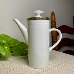 May include: A white porcelain coffee pot with a cylindrical body, a curved handle, and a tapered spout. The lid features a flat, circular knob and a decorative gold band with an intricate black scrollwork pattern around the top edge.