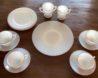 May include: A vintage white tea set with gold trim, including cups, saucers, and plates. The set features a ribbed design and is arranged on a dark wooden surface. Includes a creamer and sugar bowl.