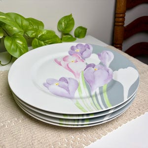 May include: A stack of three white ceramic dinner plates featuring a painted floral design of purple and pink crocus flowers with green stems. The plates have a smooth, round edge and a clean, white background with a soft grey accent area.