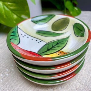 Puede incluir: Una pila de cuatro cuencos peque&ntilde;os y redondos de cer&aacute;mica. Cada cuenco presenta un interior blanco con un dise&ntilde;o pintado de hojas verdes, detalles rojos y amarillos y un borde verde. Los cuencos est&aacute;n apilados.