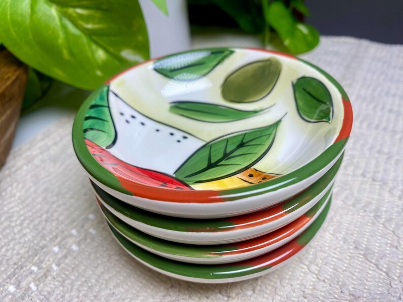 Puede incluir: Una pila de cuatro cuencos peque&ntilde;os y redondos de cer&aacute;mica. Cada cuenco presenta un interior blanco con un dise&ntilde;o pintado de hojas verdes, detalles rojos y amarillos y un borde verde. Los cuencos est&aacute;n apilados.