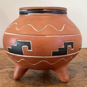 May include: A terracotta-colored ceramic pot with three short legs. The pot features black and white geometric designs and a wavy white line. The rim is dark brown. This decorative piece is likely handmade and suitable for home decor.