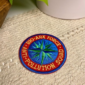 May include: Embroidered patch with a red border and the words "BIO-ARK FORCE SQUAD" in yellow. The center features a blue compass rose with green accents. The patch is circular and has a detailed design.
