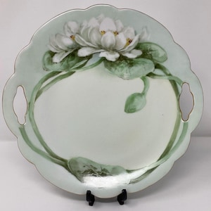 May include: A round, scalloped-edge serving plate with a pale green border and two handles. The plate features a hand-painted design of white water lilies with green leaves and stems. The plate is supported by a black stand.
