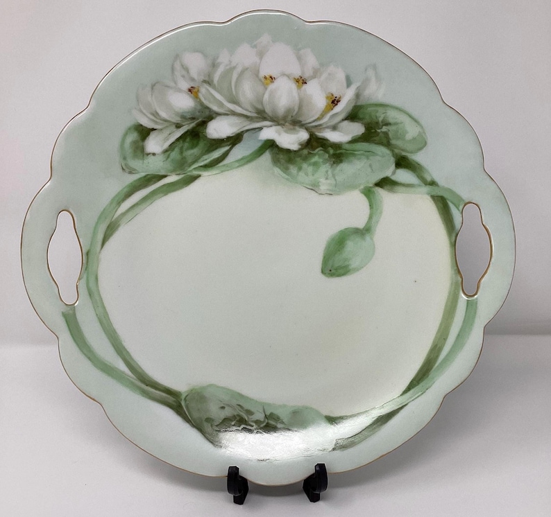 May include: A round, scalloped-edge serving plate with a pale green border and two handles. The plate features a hand-painted design of white water lilies with green leaves and stems. The plate is supported by a black stand.