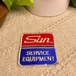 May include: A square patch with a red top section and a blue bottom section. The word "Sun" is embroidered in white on the red section. The words "SERVICE EQUIPMENT" are embroidered in white on the blue section.